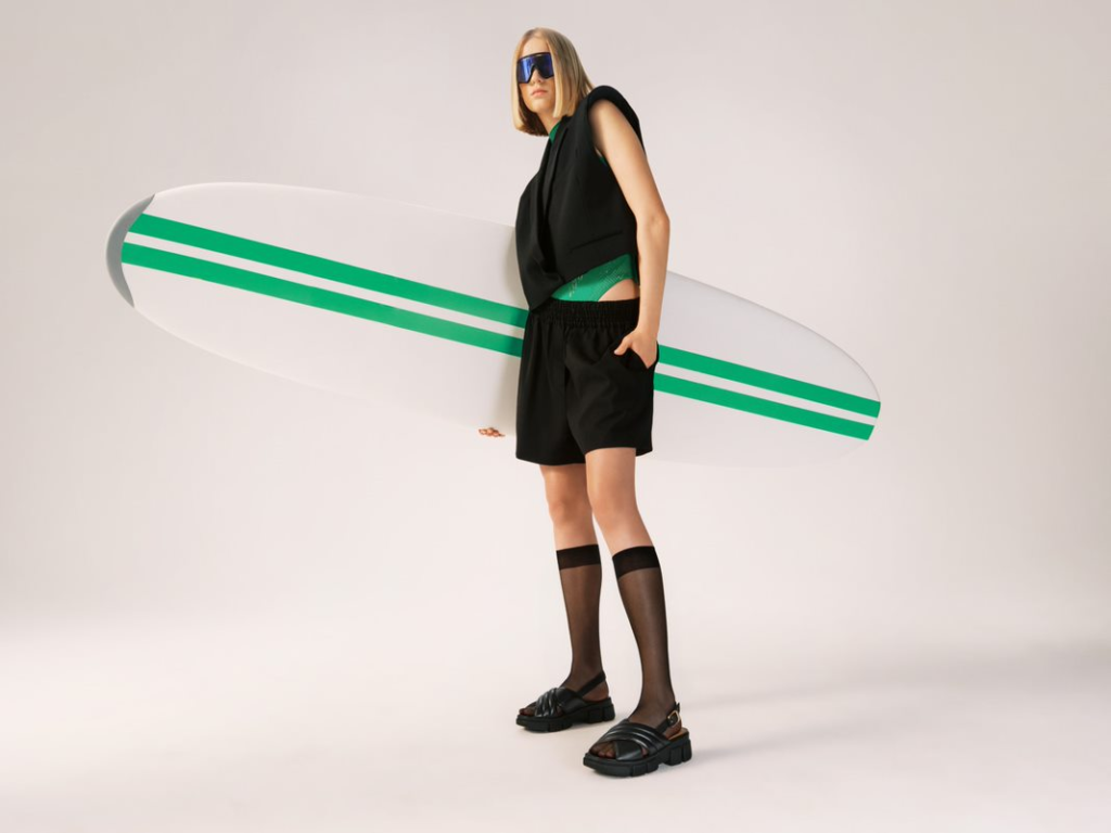 Modella in total look nero con sandali neri, tiene in mano una tavola da surf - Campagna estate 2023
